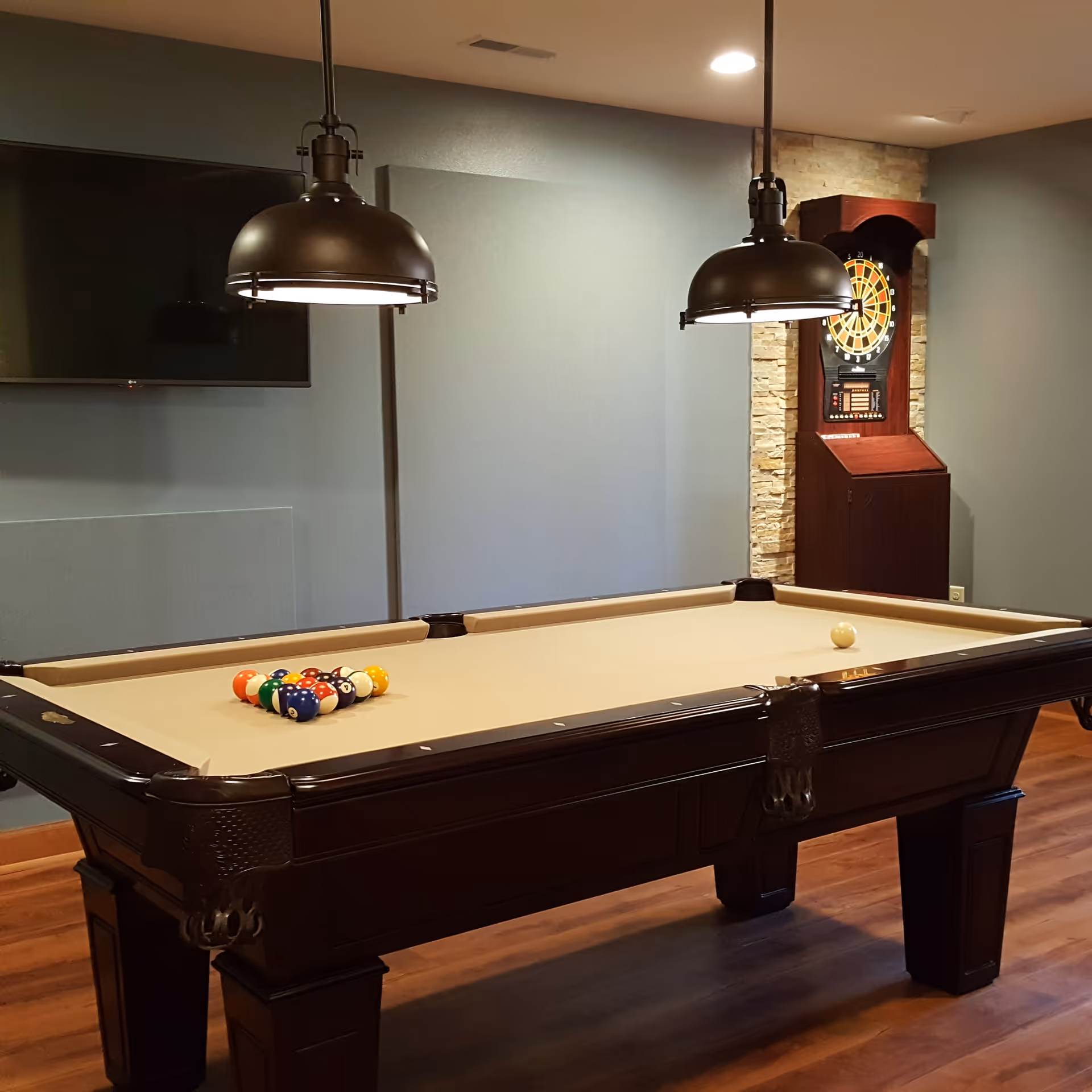Indoor recreational room with a pool table set up for a game, two hanging pendant lights above the table, a dartboard cabinet mounted on a stone accent wall, and a flat-screen TV mounted on a gray wall.