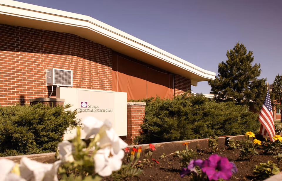 Brick senior care facility exterior with sign, flowers, shrubs, and an American flag.