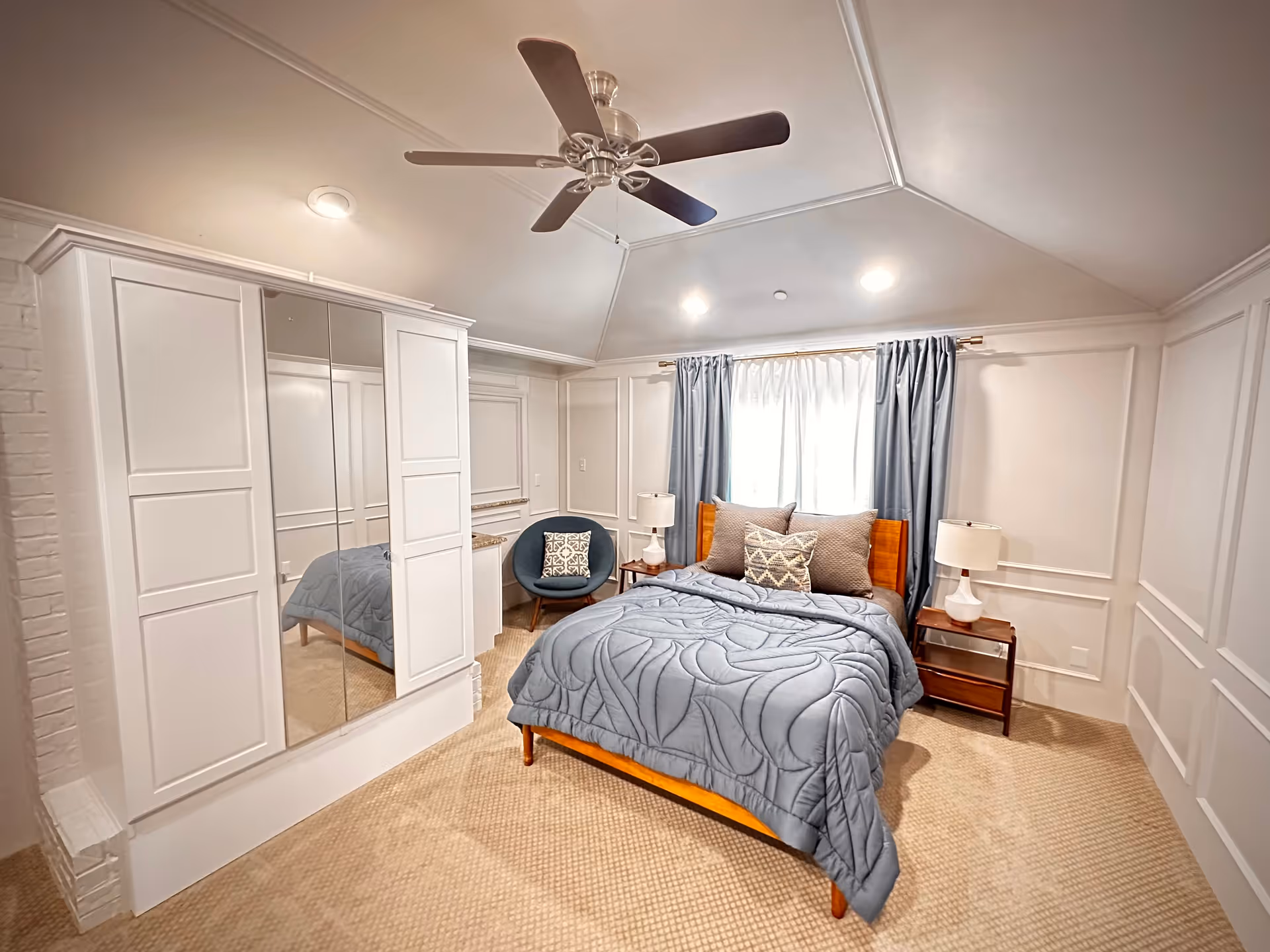 Bedroom in a senior living facility with a blue quilt, mirrored wardrobes, and ceiling fan.