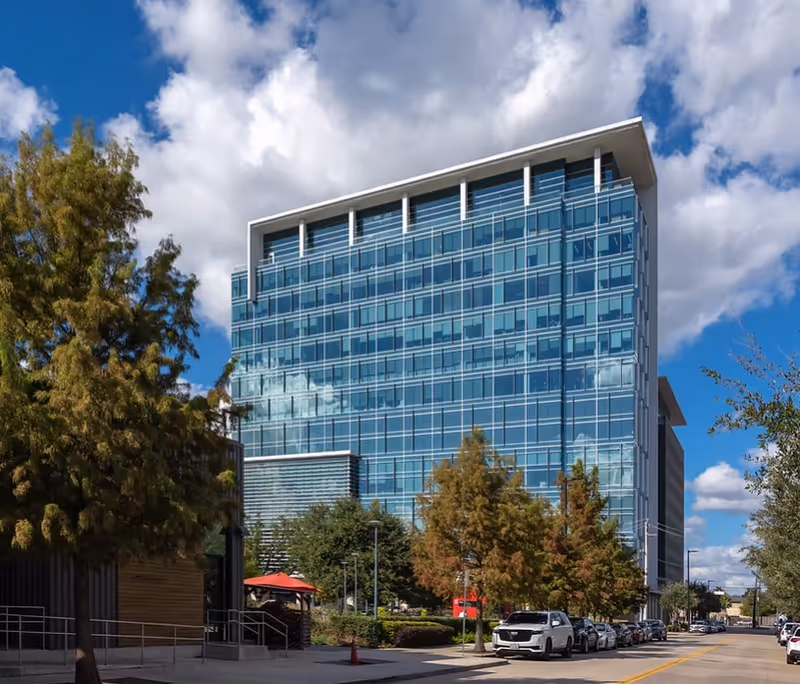 Modern glass-fronted building along a street with trees and parked cars.