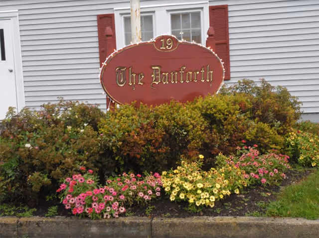 Exterior of The Danforth building with a red sign surrounded by colorful flowering shrubs.