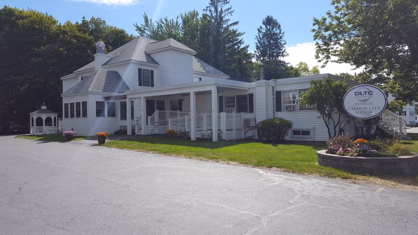White two-story senior living building with a covered porch, gazebo, landscaping, and a sign reading 'Capitol City Manor'.