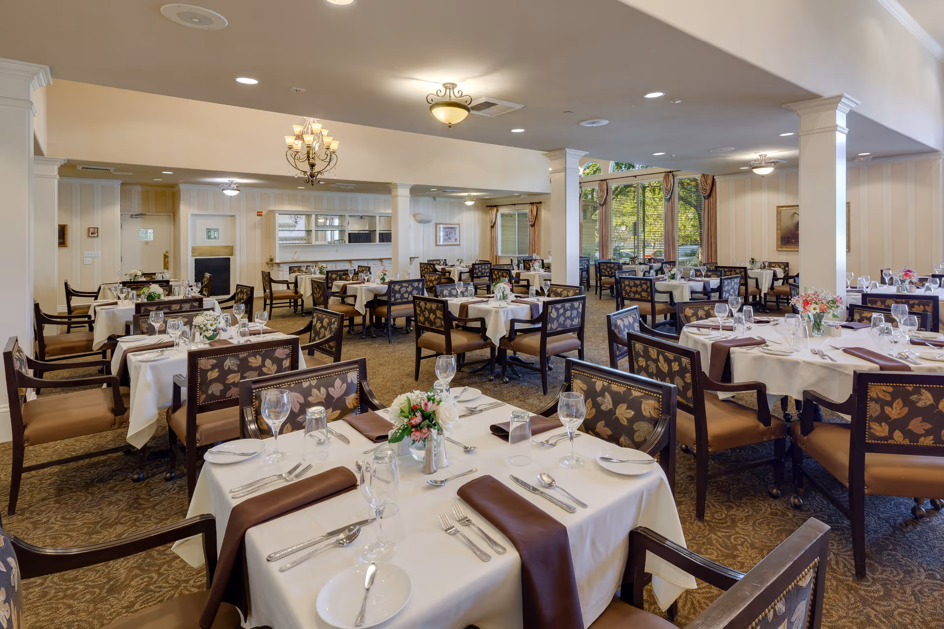A spacious dining room with multiple tables covered in white tablecloths and set with plates, silverware, glasses, and brown napkins. The chairs have patterned upholstery with a floral design. The room features beige walls, carpeted floors, large windows with curtains letting in natural light, and ceiling lights including chandeliers.