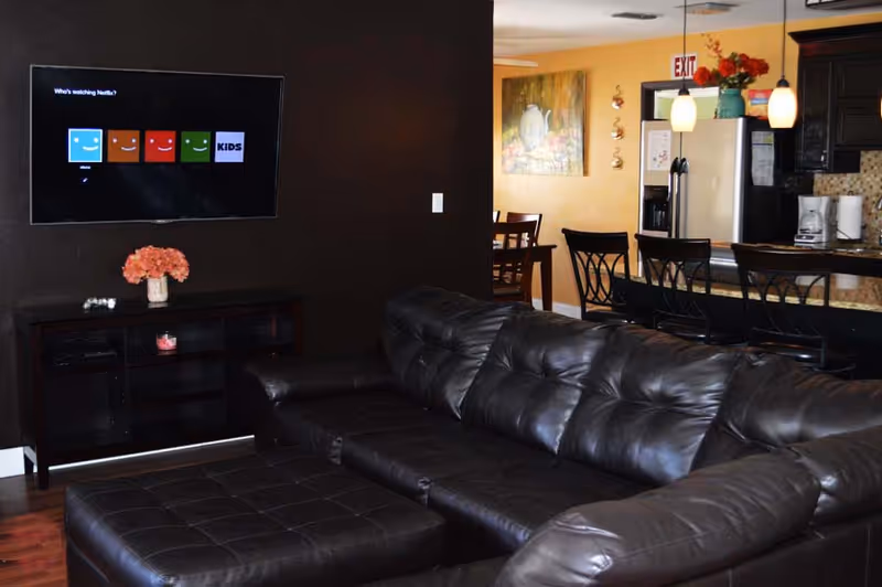 A wide view of dark living room with black leather sofa and kitchen in background.