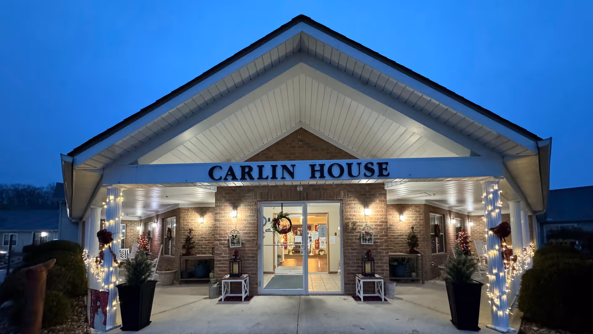 Front view of Carlin House Assisted Living entrance with brick facade and festive lights.