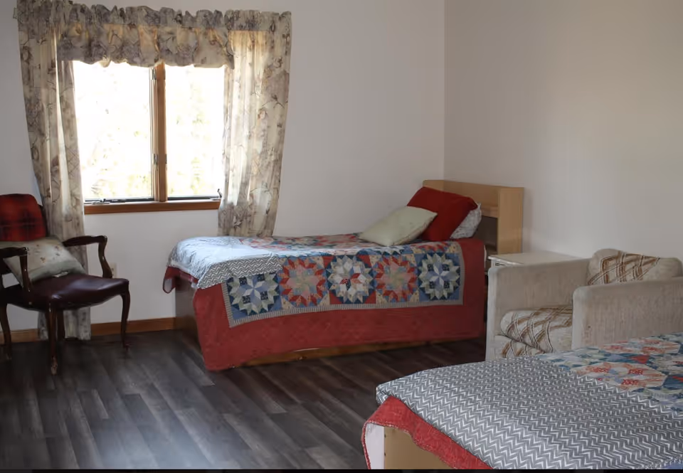 A furnished senior living bedroom with two beds, patchwork quilts, and seating by a window.