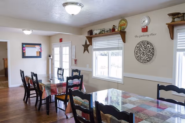 A dining area with two tables covered in colorful patchwork tablecloths and surrounded by black wooden chairs. The room has light beige walls with white trim, a window with blinds, and a glass door leading outside. Wall decorations include a clock, a metal circular wall art piece, a star, and a sign that reads 'The Gathering Place'.