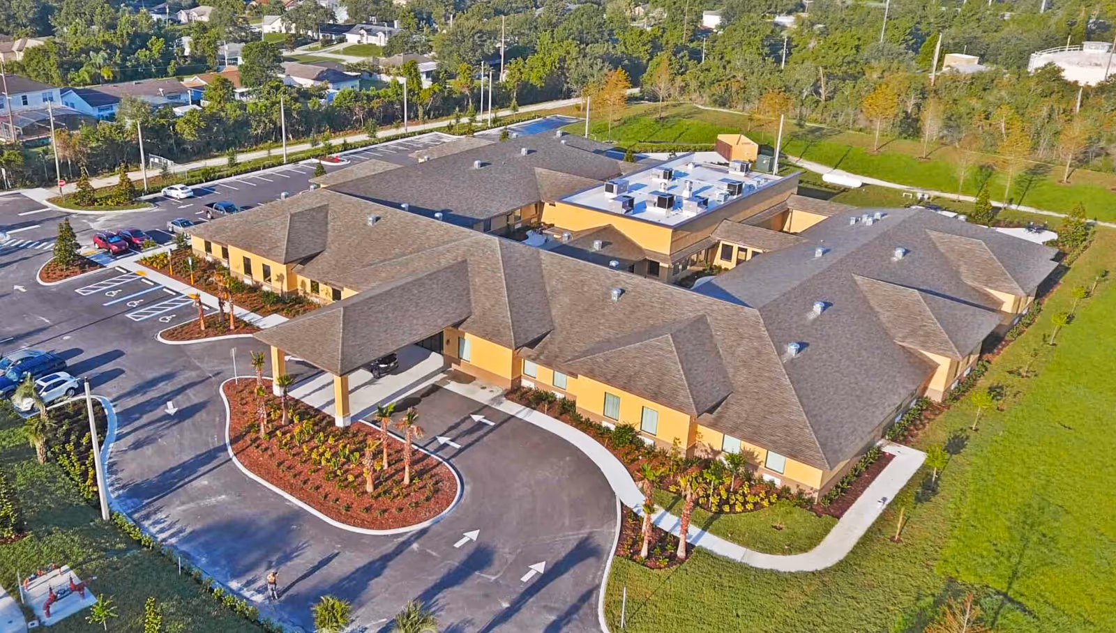 Aerial view of a single-story senior living facility with a large parking lot, landscaped entrance with palm trees, and surrounding green areas. The building has a brown roof and beige exterior walls.