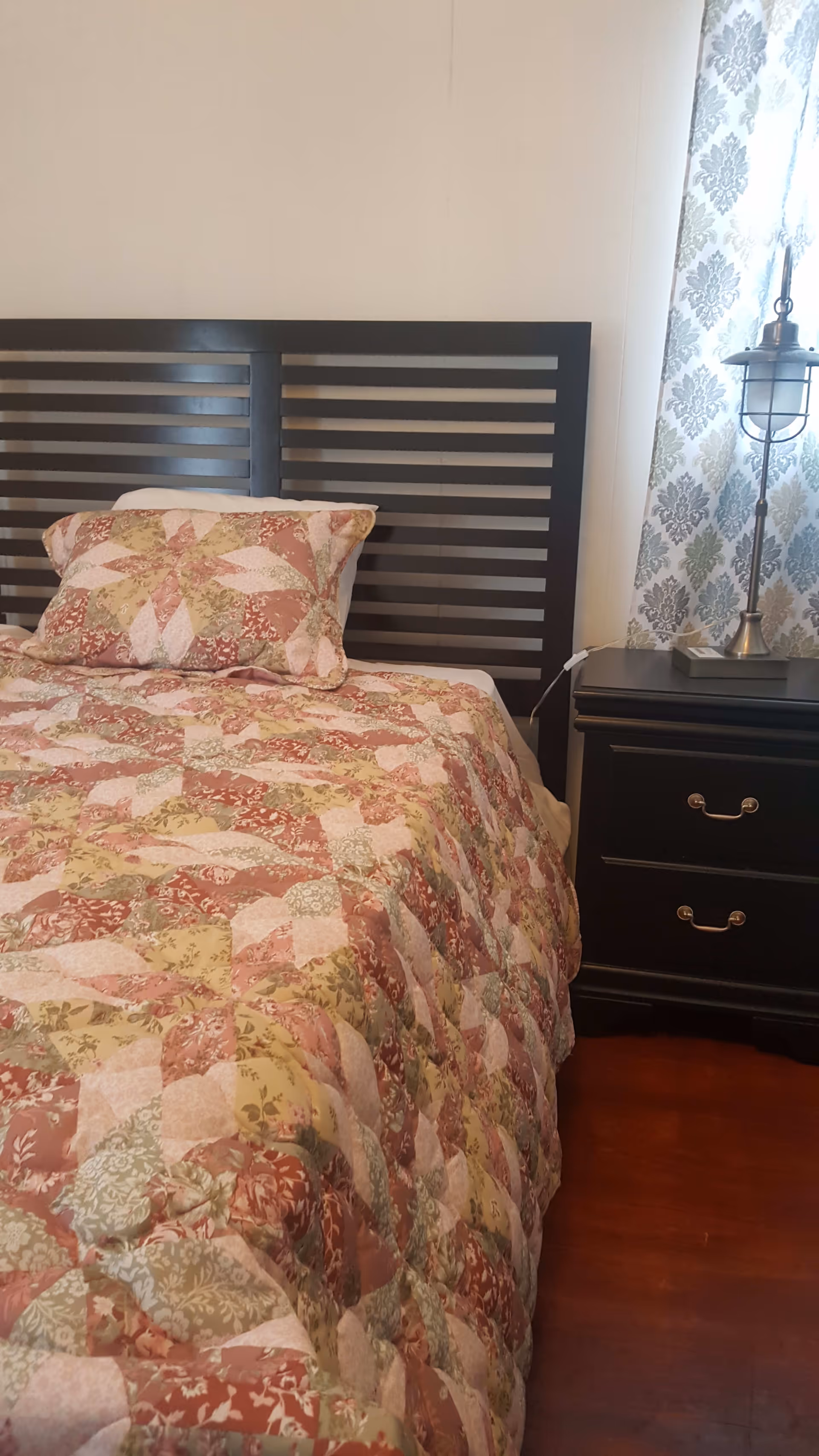 A bedroom with a bed featuring a dark wooden slatted headboard. The bed is covered with a quilted bedspread and matching pillow in a patchwork pattern of pink, green, and cream colors. Next to the bed is a dark wooden nightstand with two drawers and a decorative lamp with a metal cage shade. A window with patterned curtains is partially visible behind the nightstand.
