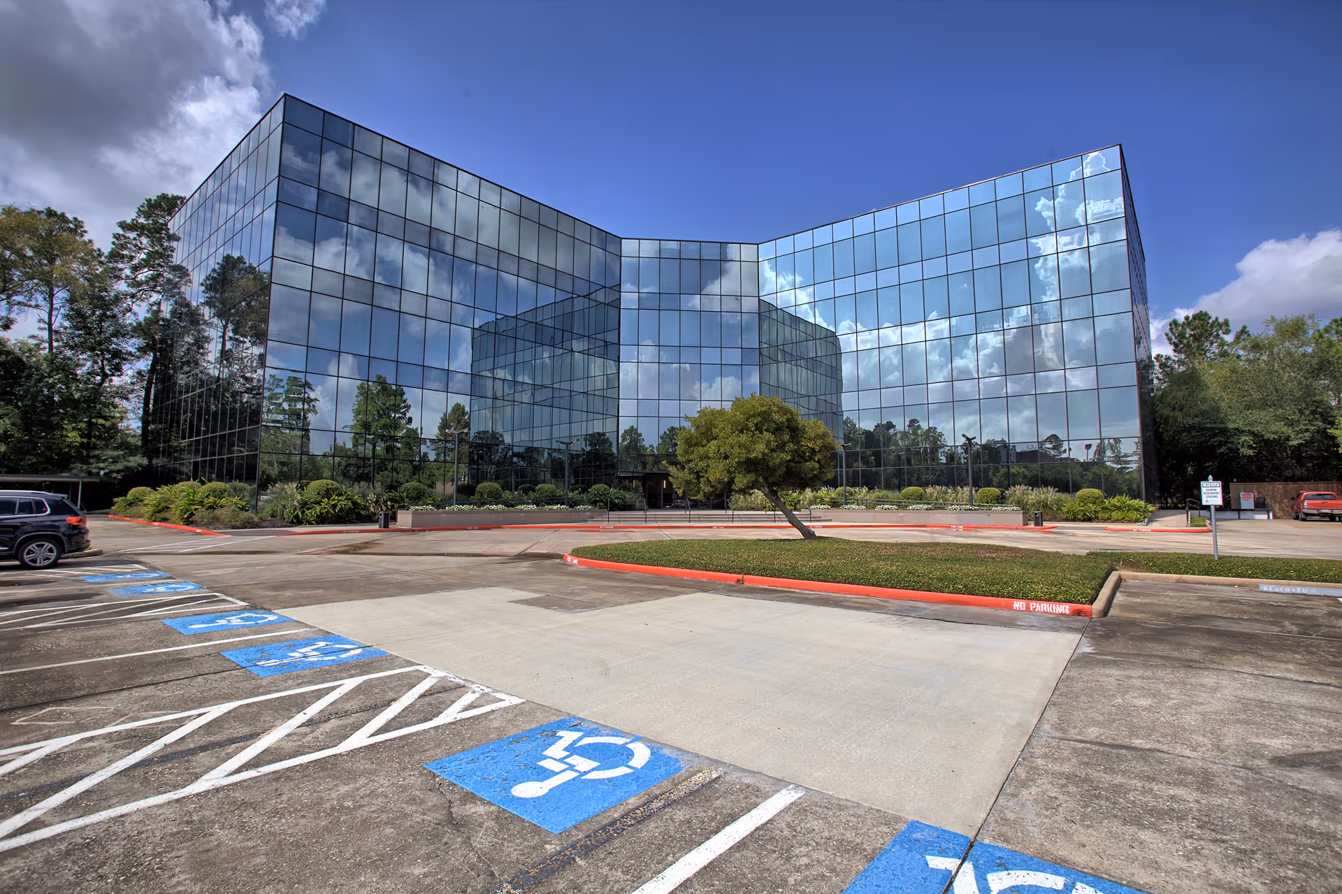 Glass-front office building reflects clouds, with a parking lot and landscaped entrance.