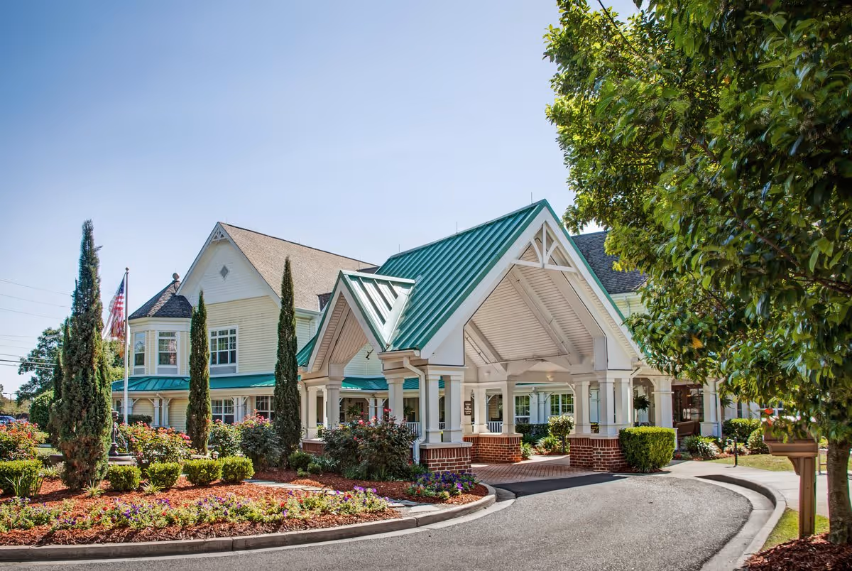 Front entrance of a senior living facility with a green metal roof, covered porte-cochère, landscaped flower beds, and a circular driveway.