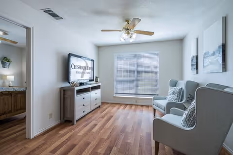 Bright living room with two gray armchairs, a TV stand, large window, and wooden floors.