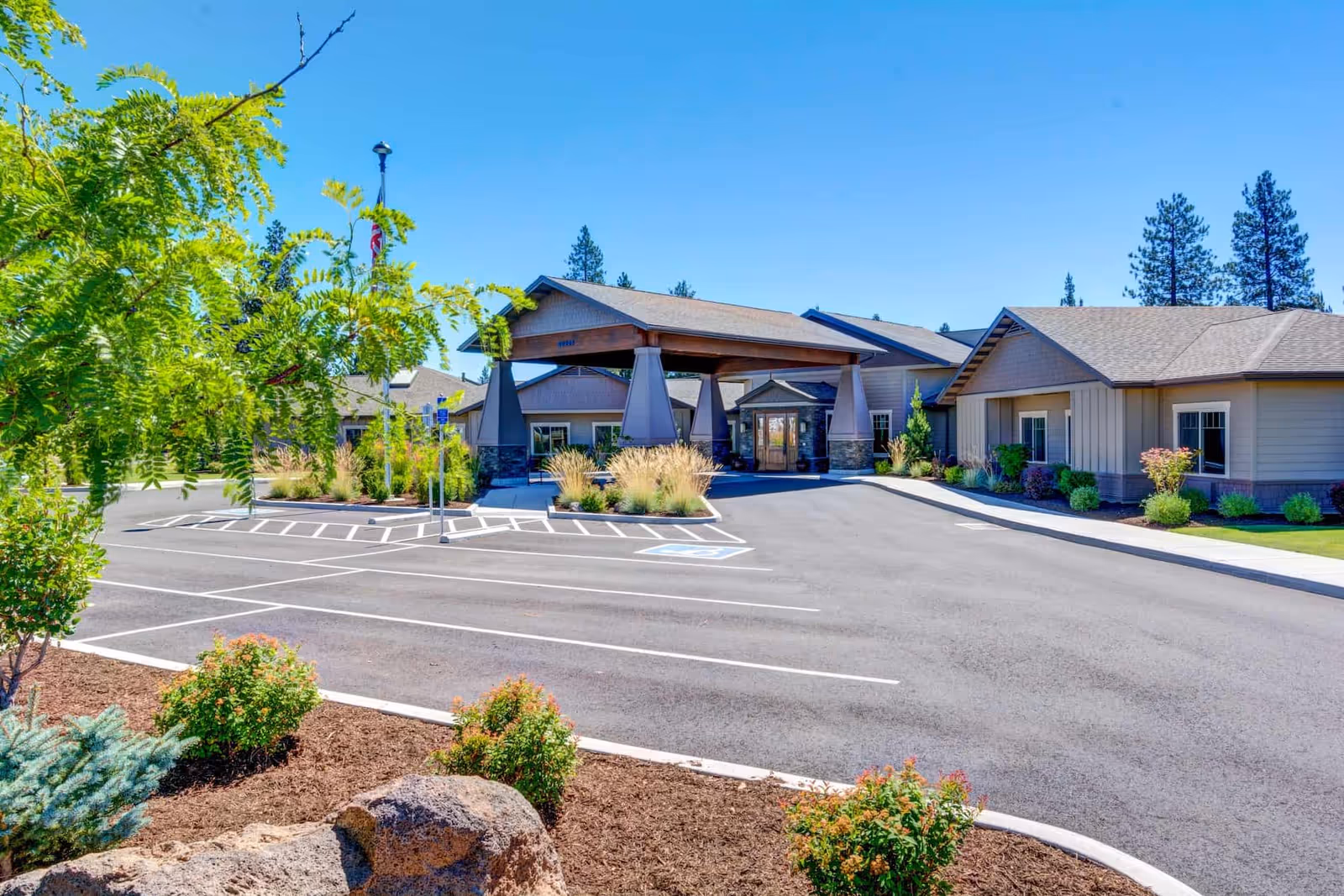Front entrance of Mt. Bachelor Assisted Living and Memory Care with a covered porte-cochere, parking lot, and landscaped grounds under a clear blue sky.