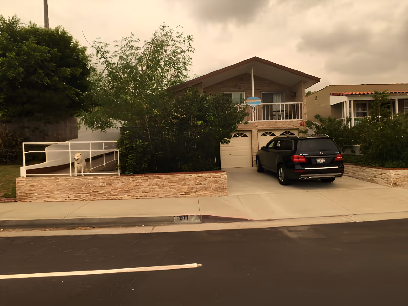 Exterior view of a two-story house with a driveway and parked black SUV, garage doors, balcony, greenery, and a dog behind a front railing.