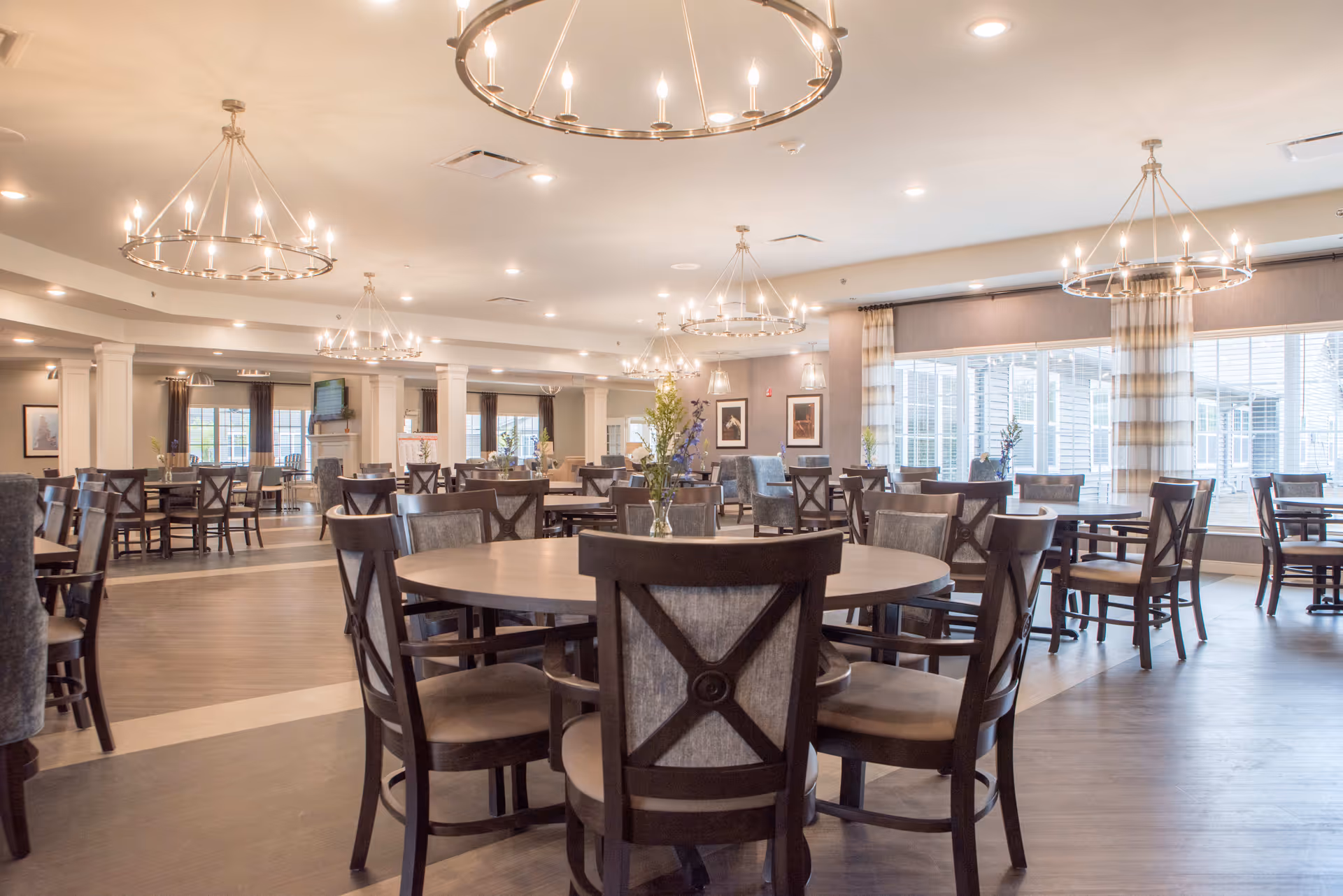 Spacious senior living dining room with round wooden tables, cushioned chairs, large windows and chandelier lighting.