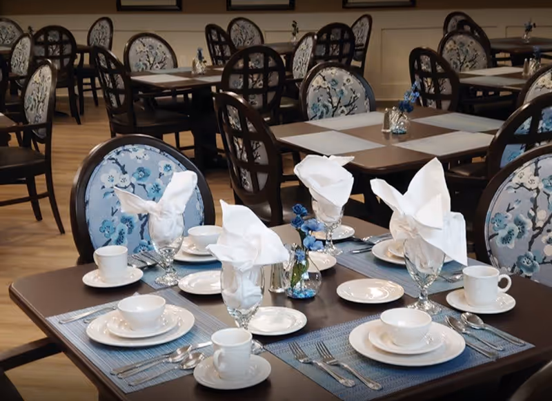 A dining room with neatly set tables, white napkin-folded glasses, place settings, and floral-upholstered chairs.