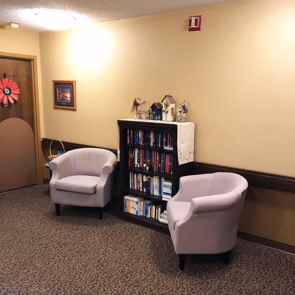 A cozy corner in an interior room featuring two light purple armchairs positioned on either side of a dark wooden bookshelf filled with books. The bookshelf has decorative birdhouses and small figurines on top. The walls are painted beige with a framed picture hanging and a door with a pink flower decoration. The floor is carpeted with a patterned design.