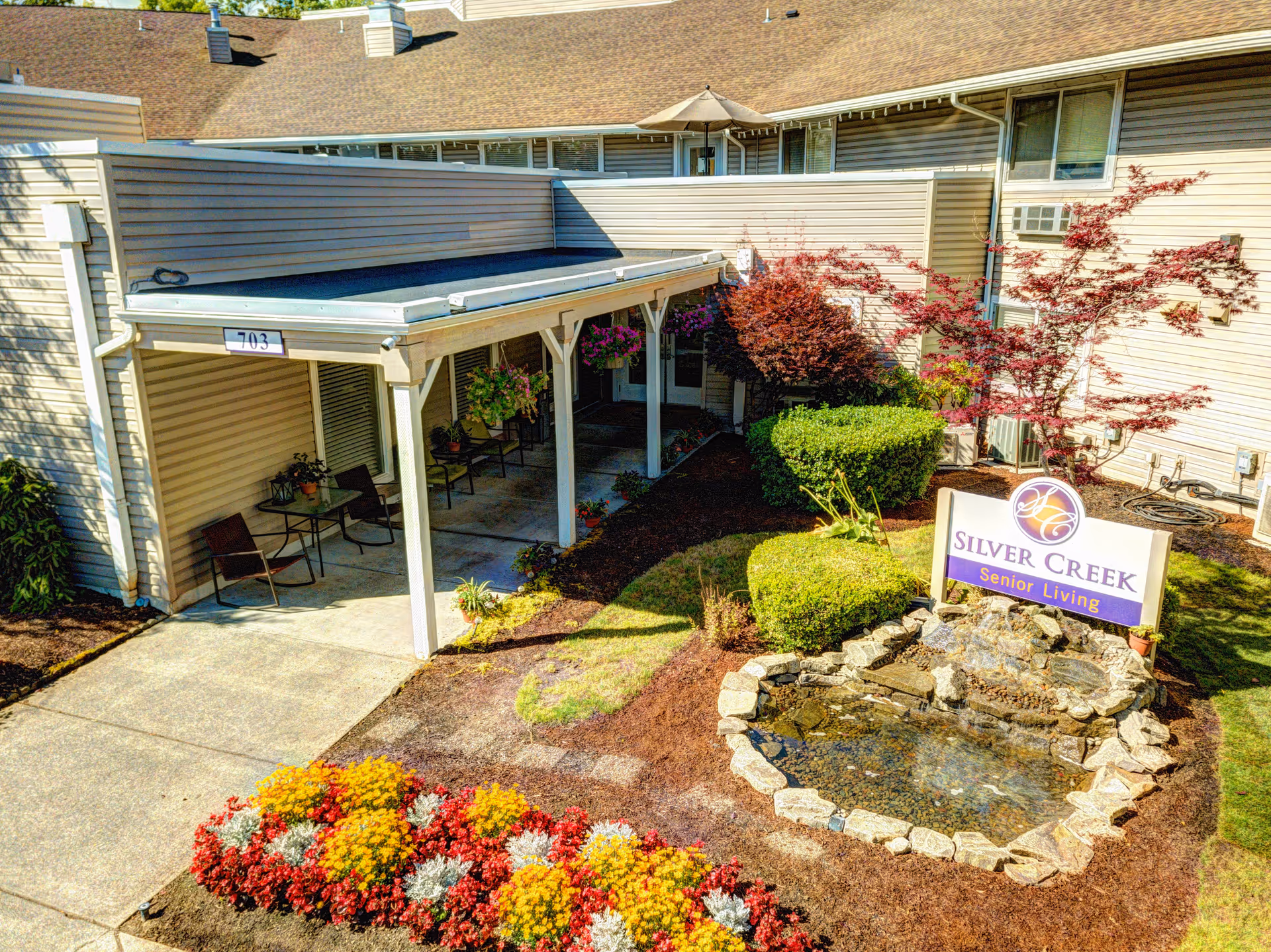 Exterior view of Silver Creek Senior Living facility showing a covered patio area with chairs and tables, a landscaped garden with colorful flowers, bushes, a small pond with rocks, and a sign that reads 'Silver Creek Senior Living'.