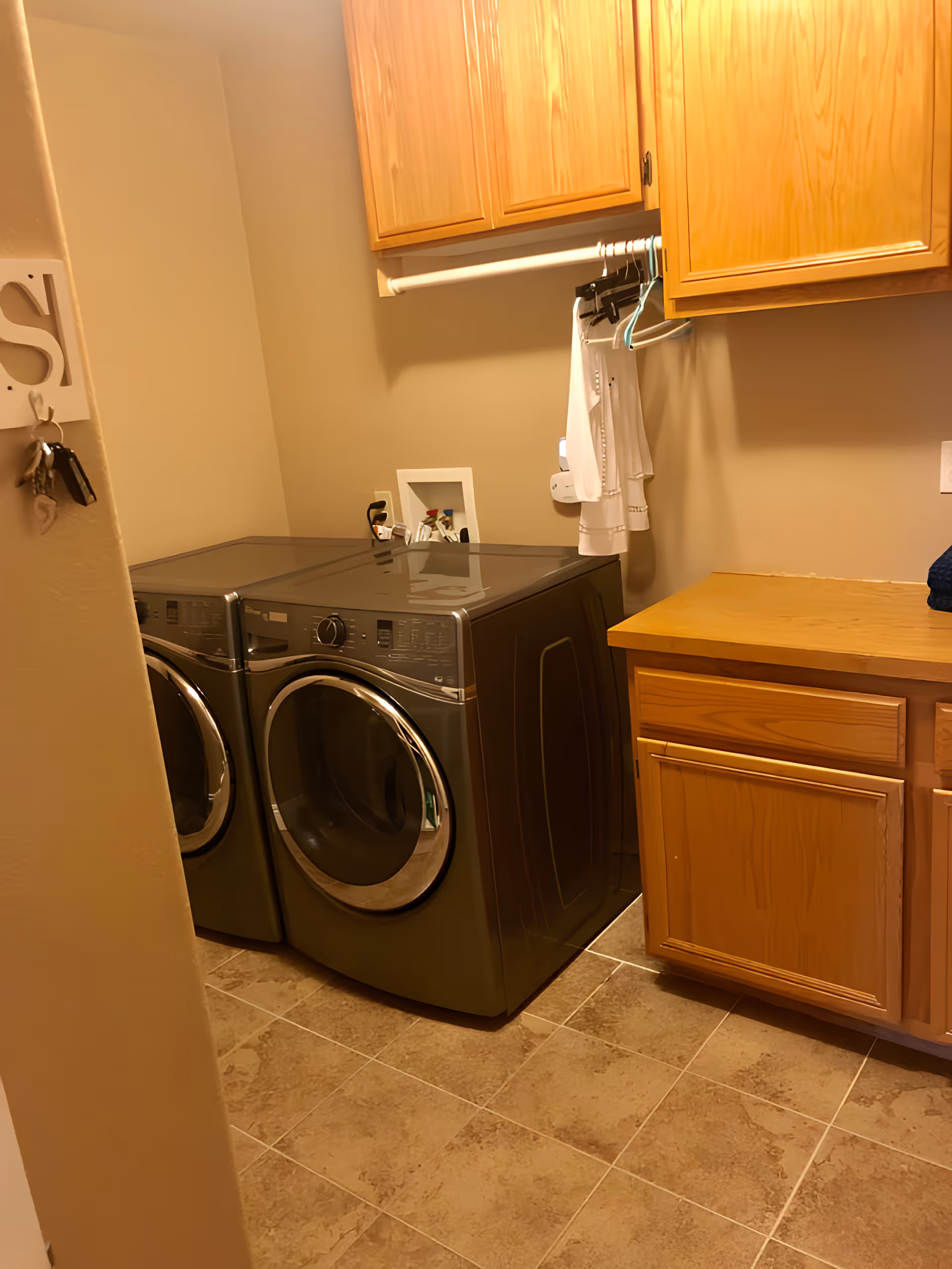 Laundry room with front-loading washer and dryer, wooden cabinets, countertop and a few hanging clothes.