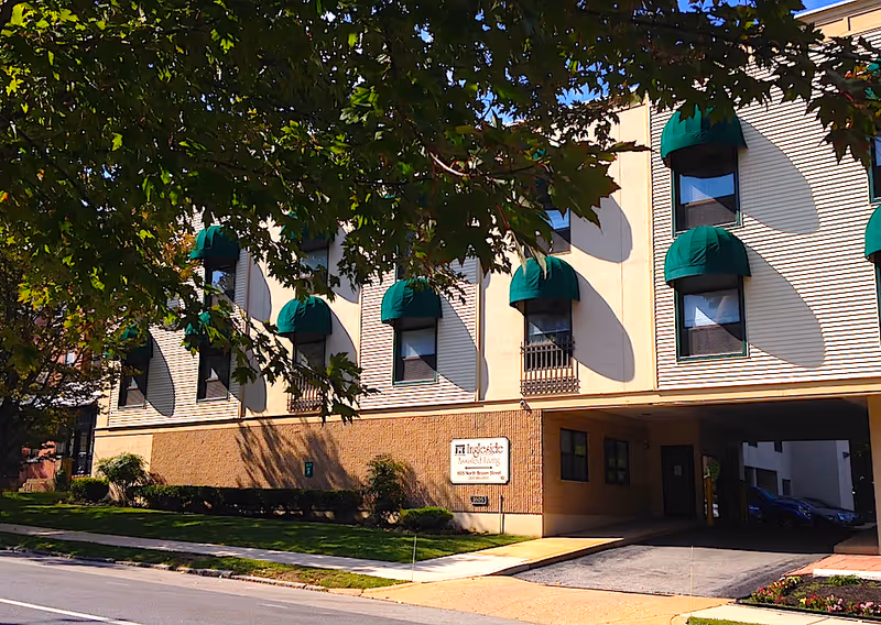 Exterior view of Ingleside Homes Assisted Living LLC building with beige walls and green awnings over the windows. There is a driveway entrance on the right side and a sidewalk with grass and small bushes in front. Tree branches with green leaves partially frame the top of the image.