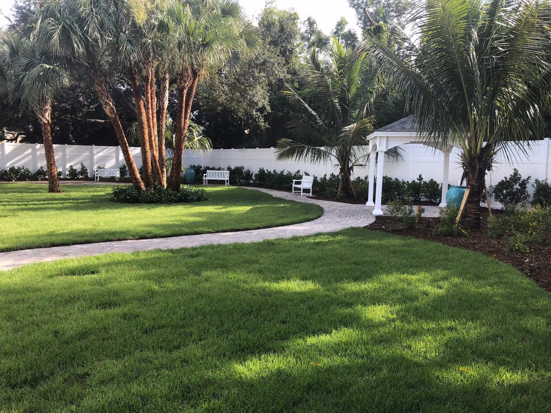 A well-maintained outdoor garden area with lush green grass, several palm trees, a curved paved walkway, white benches, and a small white gazebo. The area is enclosed by a white fence with shrubs planted along the fence line.