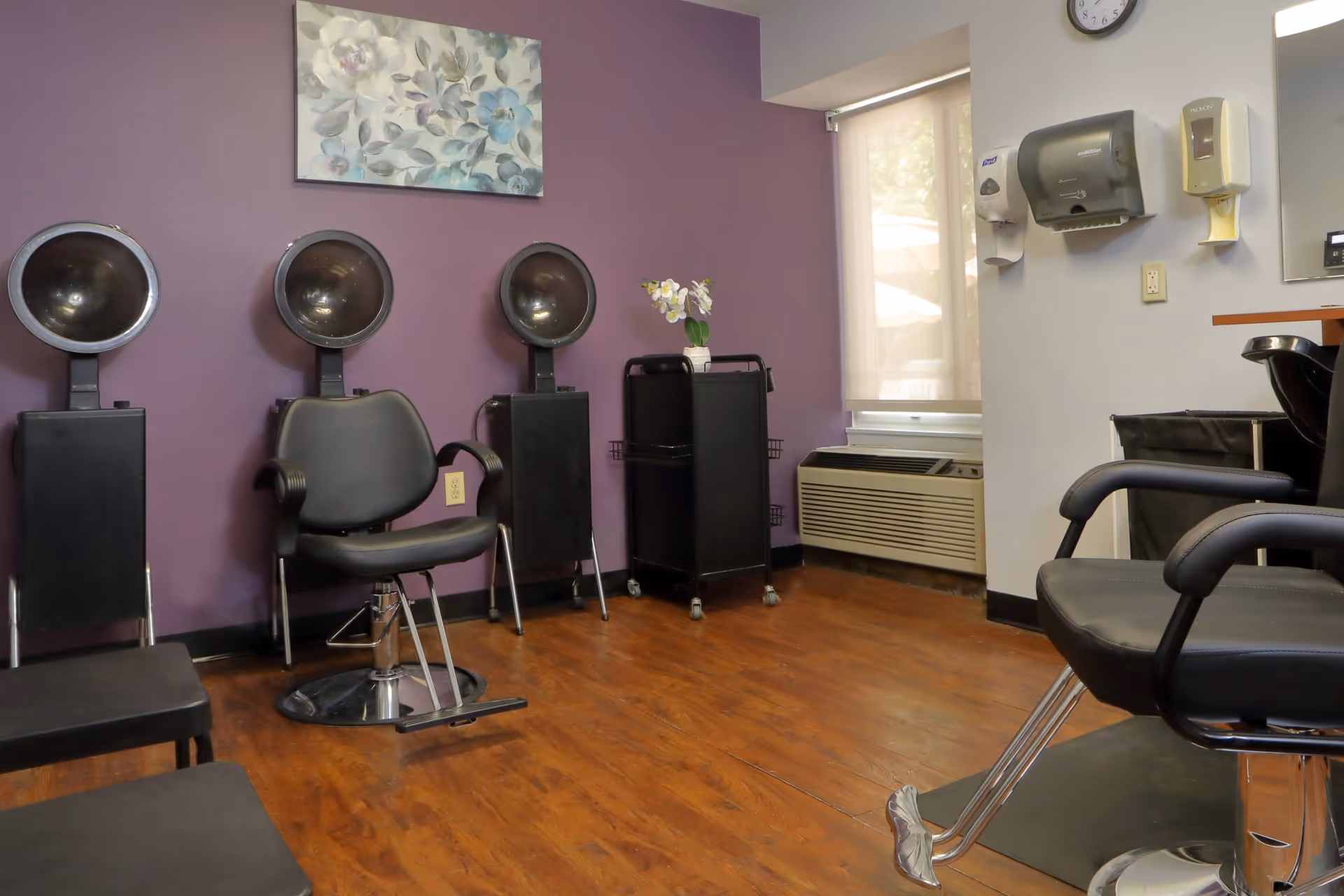 Interior of a hair salon area with three black hair dryer chairs against a purple wall, a black salon chair in the foreground, a window with a beige shade, and a wall-mounted clock and dispensers.