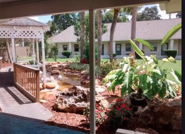 Landscaped courtyard with a wooden gazebo, rock-lined pond, tropical plants and a single-story building in the background.