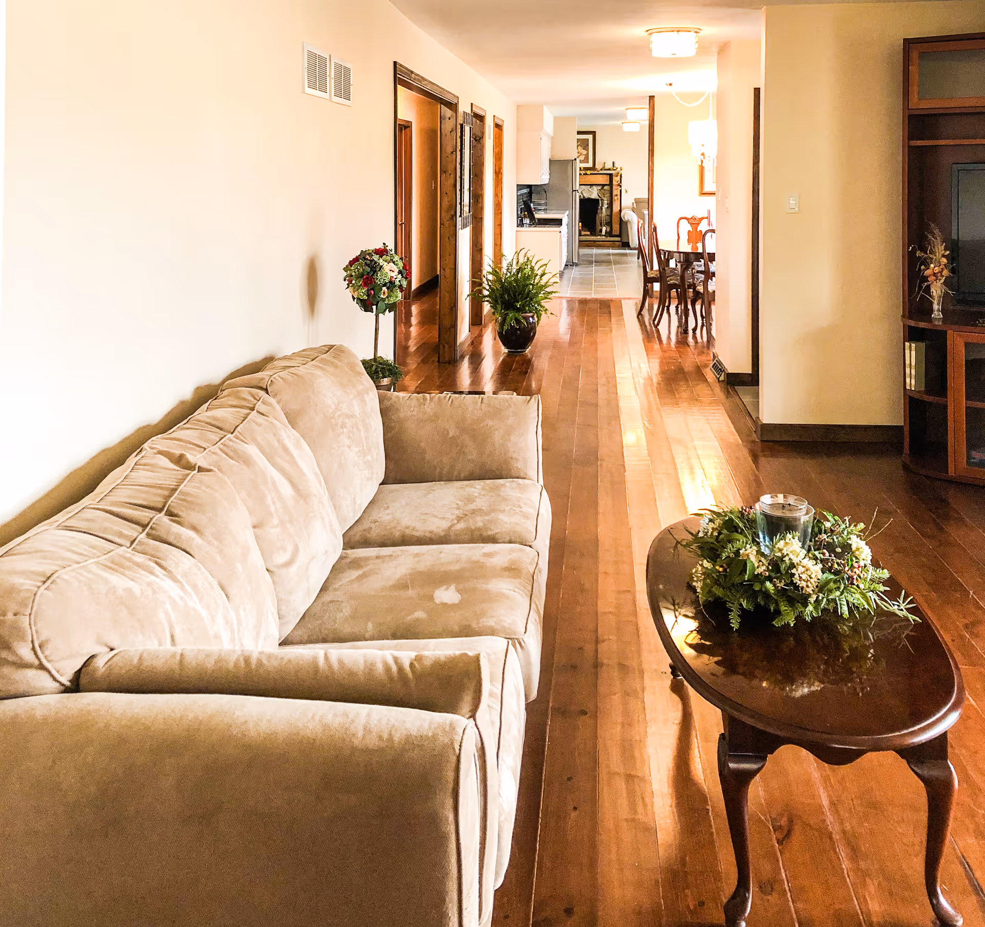 Interior view of a senior living facility showing a beige couch on the left, a wooden coffee table with a floral centerpiece and candle, hardwood floors, and a hallway leading to a dining area and kitchen in the background.