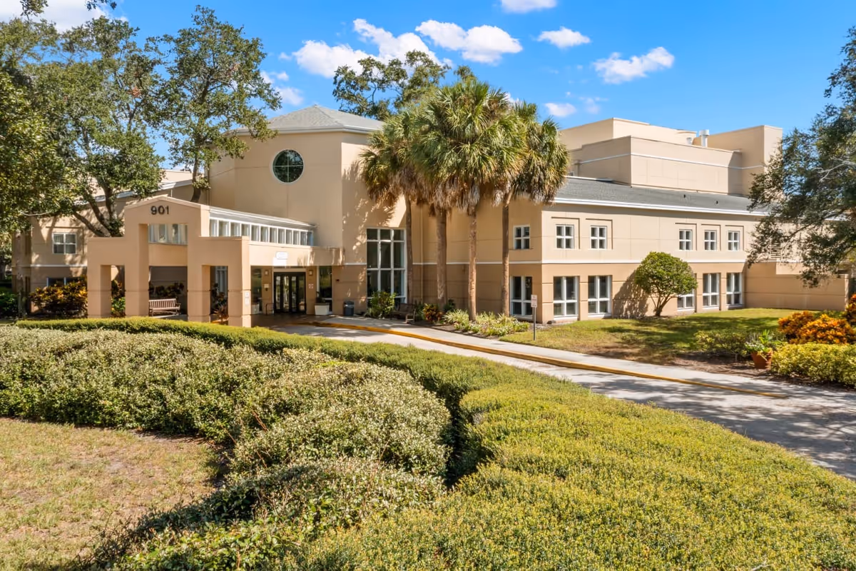 Beige senior living facility front with palm trees, trimmed hedges, and walkway entry.