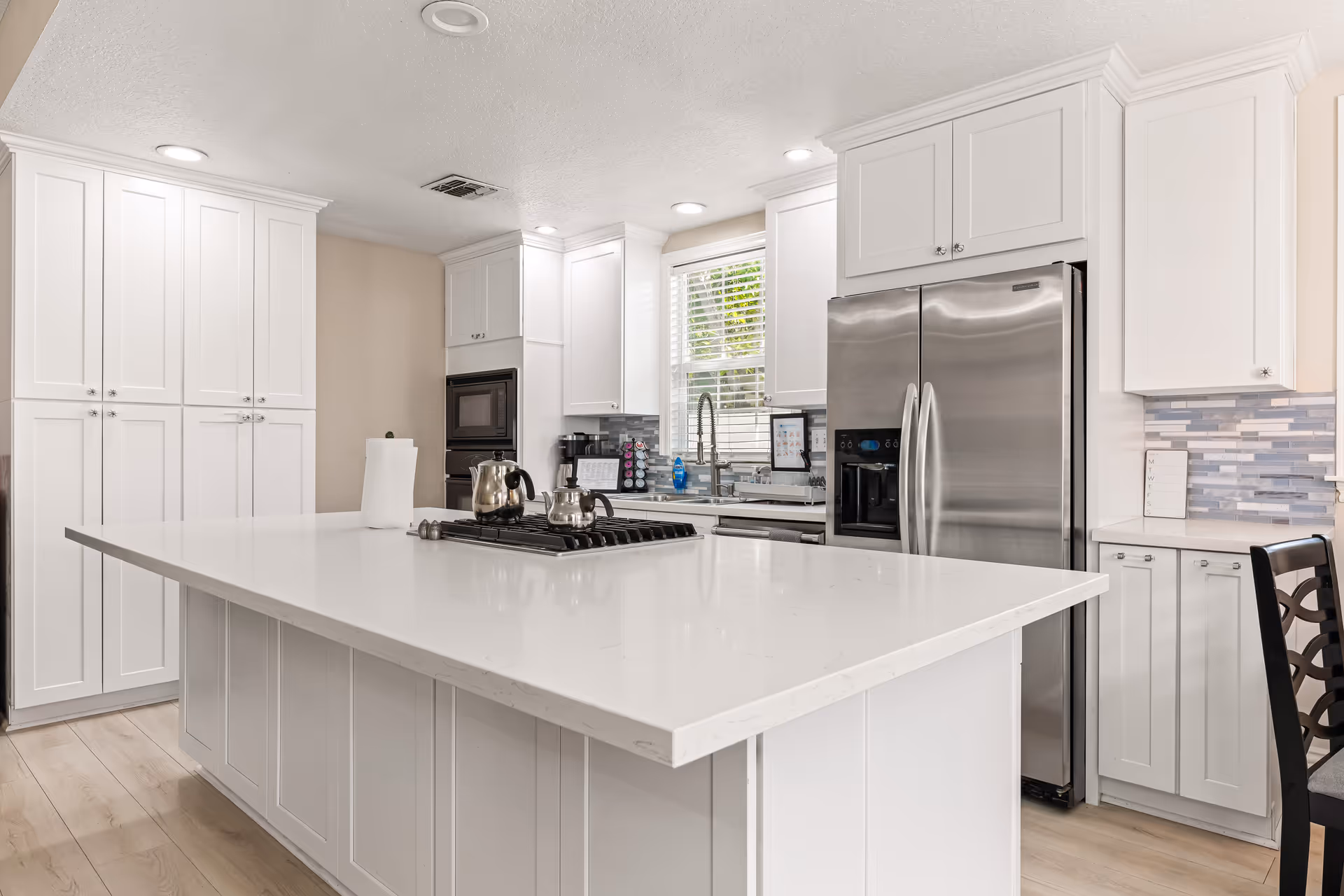 Bright white kitchen with large island, stainless steel fridge, cabinetry, and tiled backsplash.