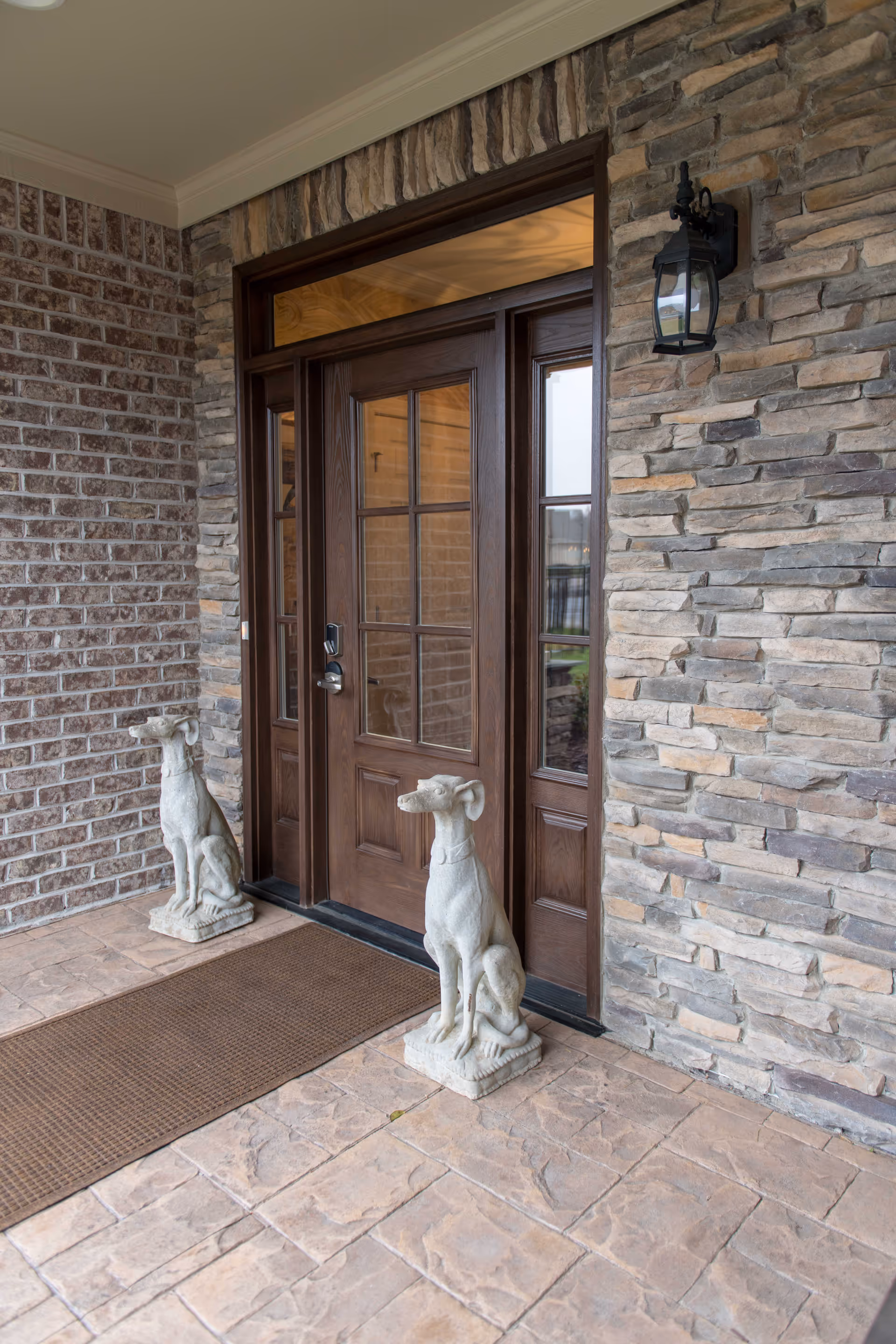 Front entrance of a building with a wooden door featuring glass panels, flanked by two stone dog statues on either side of a brown doormat. The exterior walls are made of brick and stone, and there is a black lantern-style light fixture mounted on the right side.
