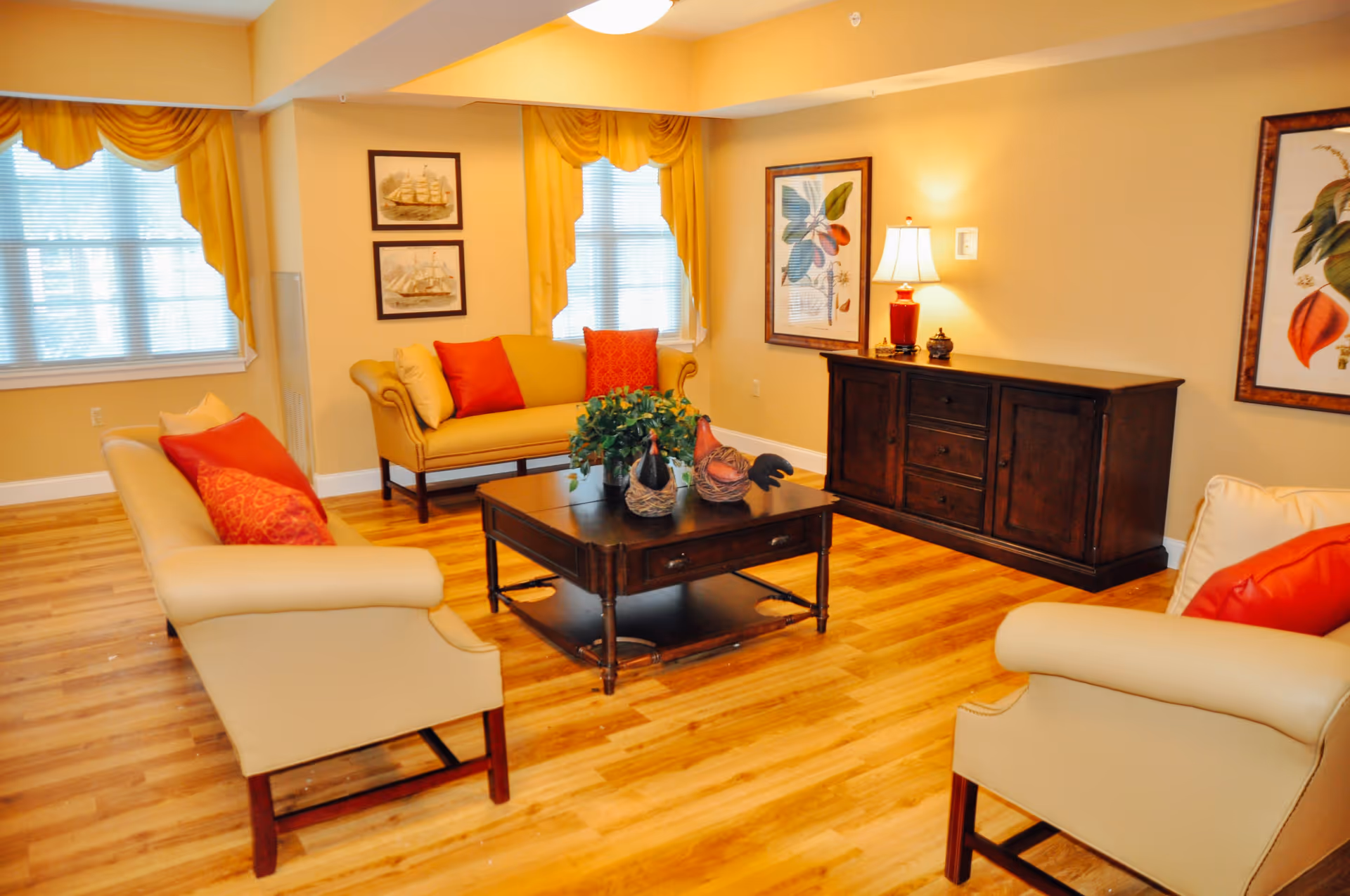 A cozy living room with wooden flooring, beige walls, and yellow curtains. The room features two beige armchairs and a yellow loveseat, all adorned with red and beige cushions. A dark wooden coffee table with decorative items and a green plant sits in the center. On the right side, there is a dark wooden sideboard with a red lamp and framed botanical artwork on the walls.