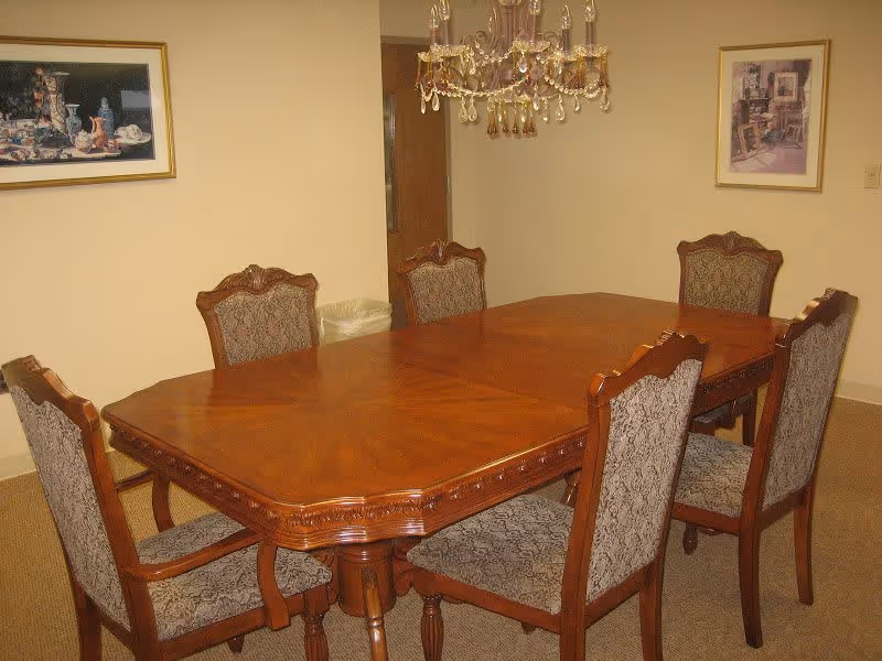 A dining room with a wooden rectangular table surrounded by six upholstered chairs with patterned fabric. A chandelier hangs above the table. Two framed pictures are on the beige walls.