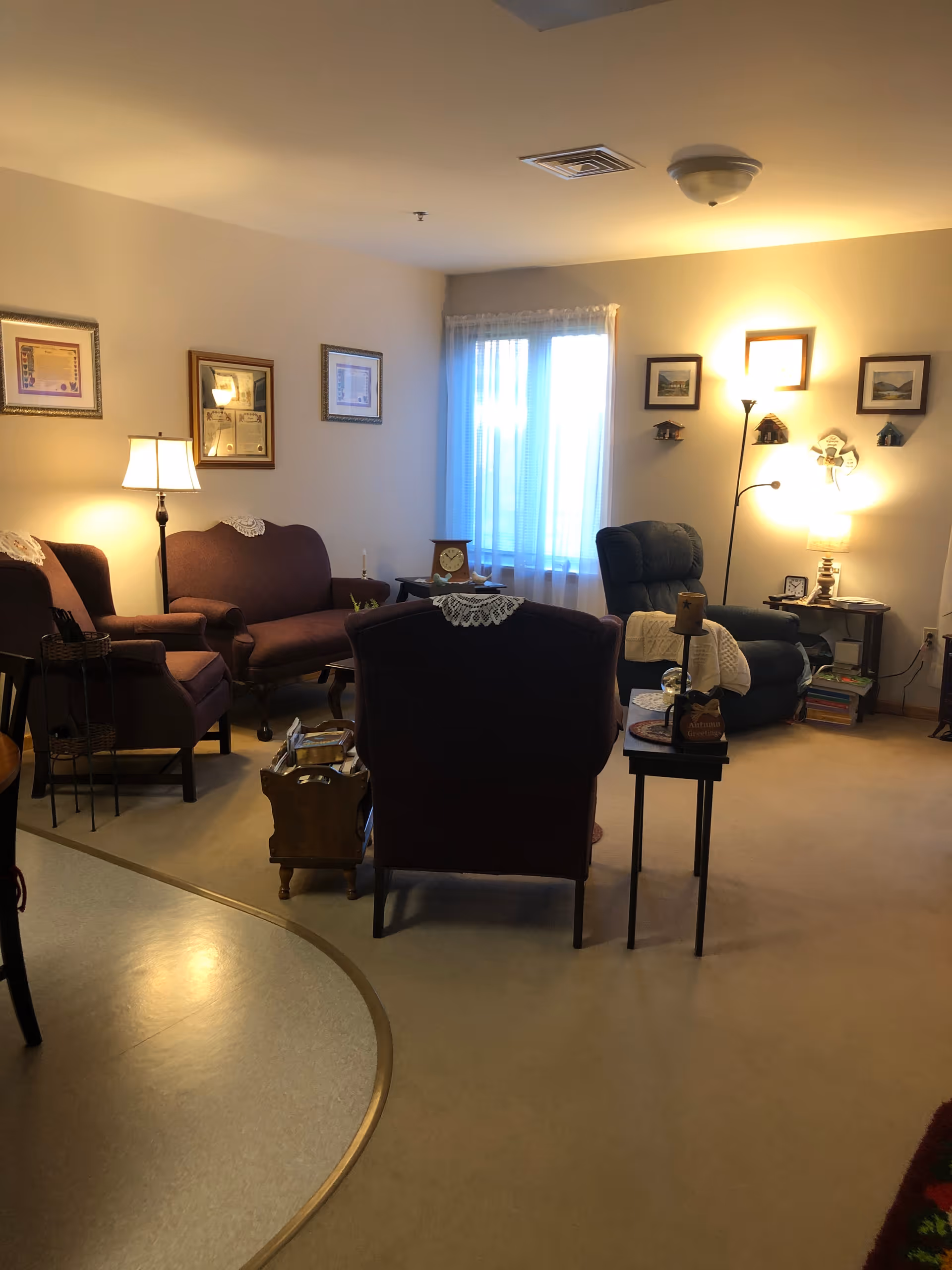 A cozy living room area with several upholstered chairs and a loveseat arranged around small tables. The room is softly lit by floor and table lamps. There are framed pictures and certificates on the walls, lace doilies on the furniture, and a window with sheer curtains letting in natural light.