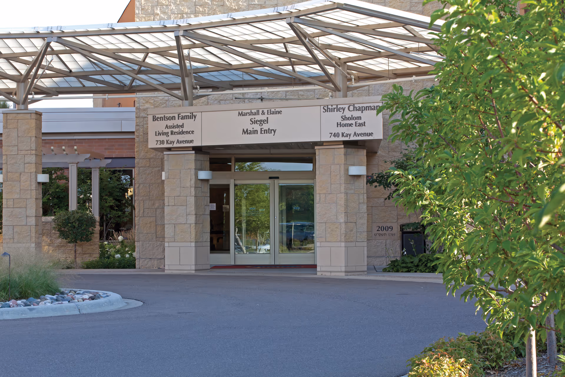 Entrance to a senior living facility with a covered driveway and glass double doors. Signs above the entrance indicate Bentson Family Assisted Living Residence at 730 Kay Avenue, Marshall & Elaine Siegel Main Entry, and Shirley Chapman Sholom Home East at 740 Kay Avenue. There are stone pillars and greenery around the entrance.