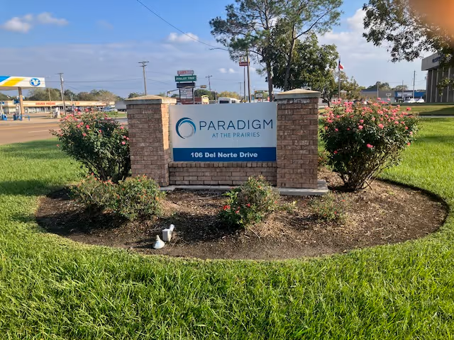 Brick entrance sign reading 'Paradigm at the Prairies, 106 Del Norte Drive' surrounded by grass and flowering shrubs with a street and businesses in the background.