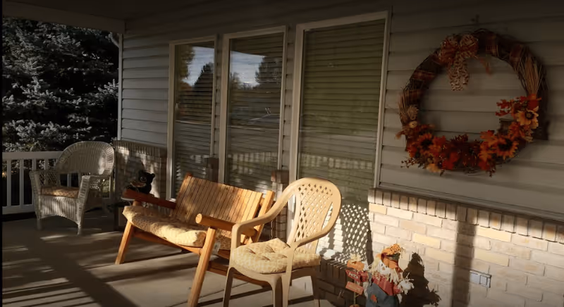 A covered outdoor porch area with a wooden bench, a plastic chair with a cushion, and a wicker chair. There is a decorative autumn wreath hanging on the wall and a small scarecrow decoration on the ground. Trees and a railing are visible in the background.