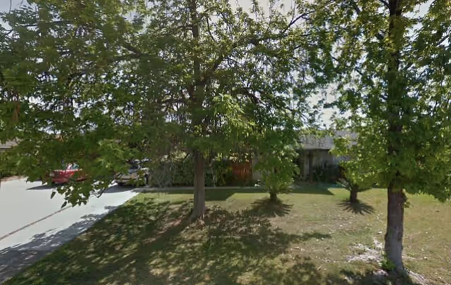 Front exterior of a senior care center partially obscured by green trees and a driveway.