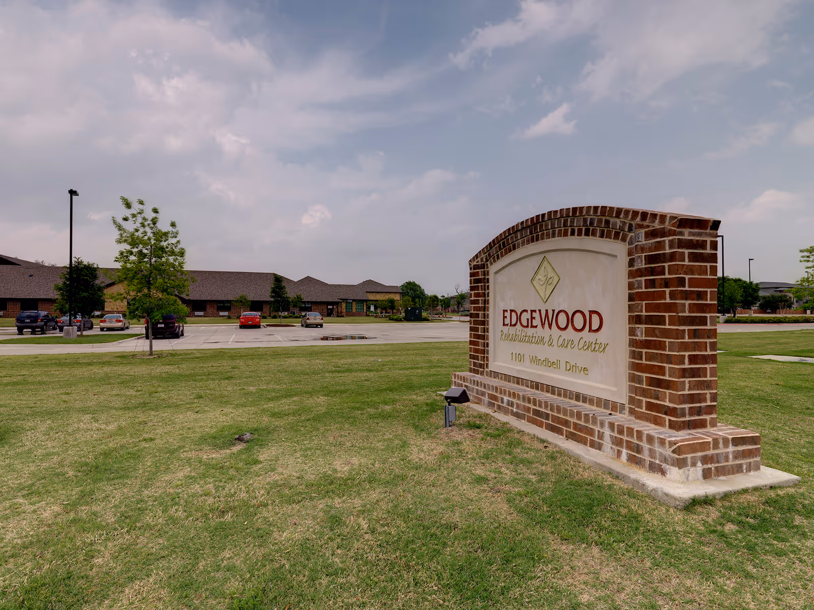 Brick entrance sign for Edgewood Rehabilitation & Care Center on a grassy lawn with the facility building and parking lot in the background.