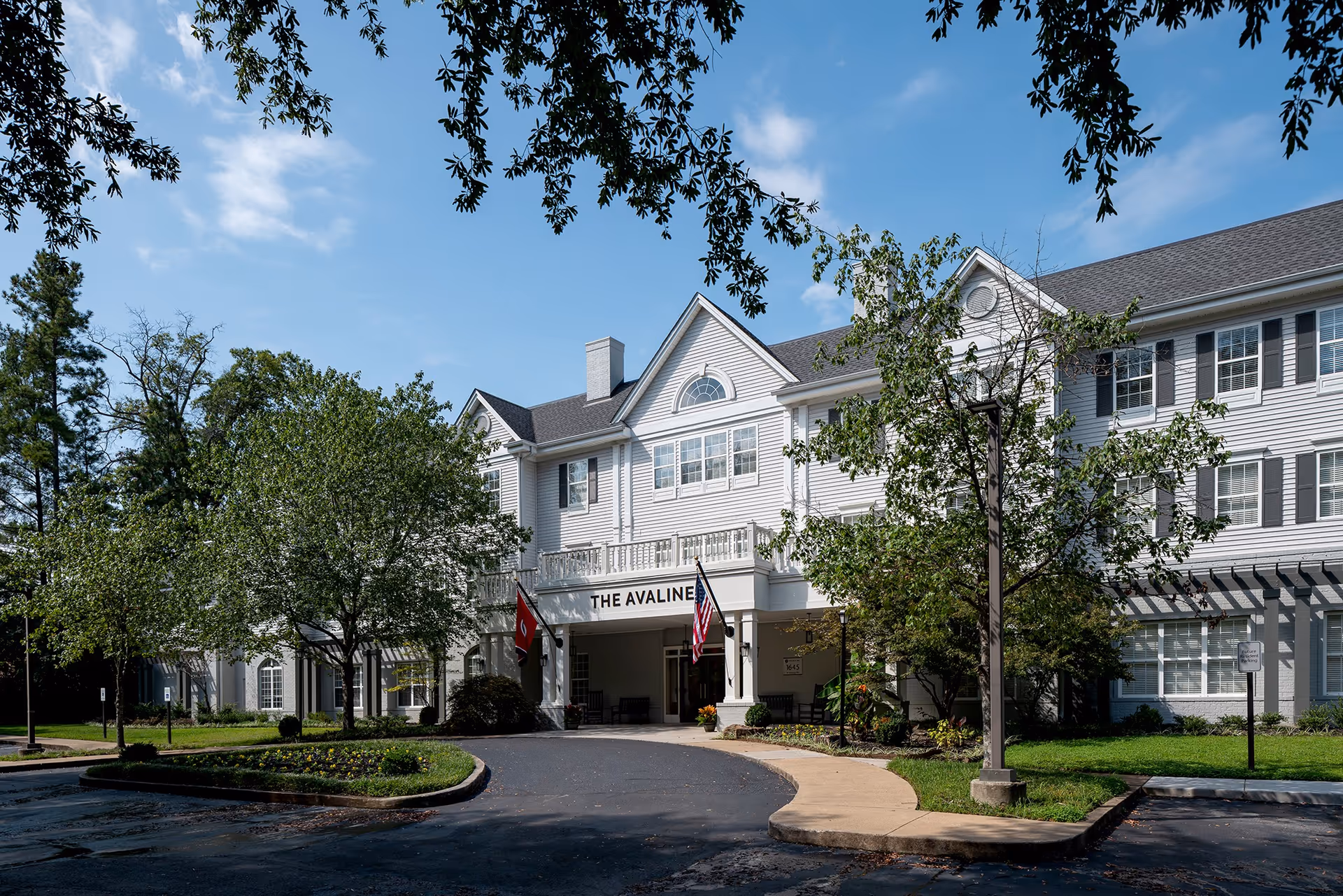 White multi-story senior living building with trees, driveway, and entrance labeled The Avaline at River Oaks.