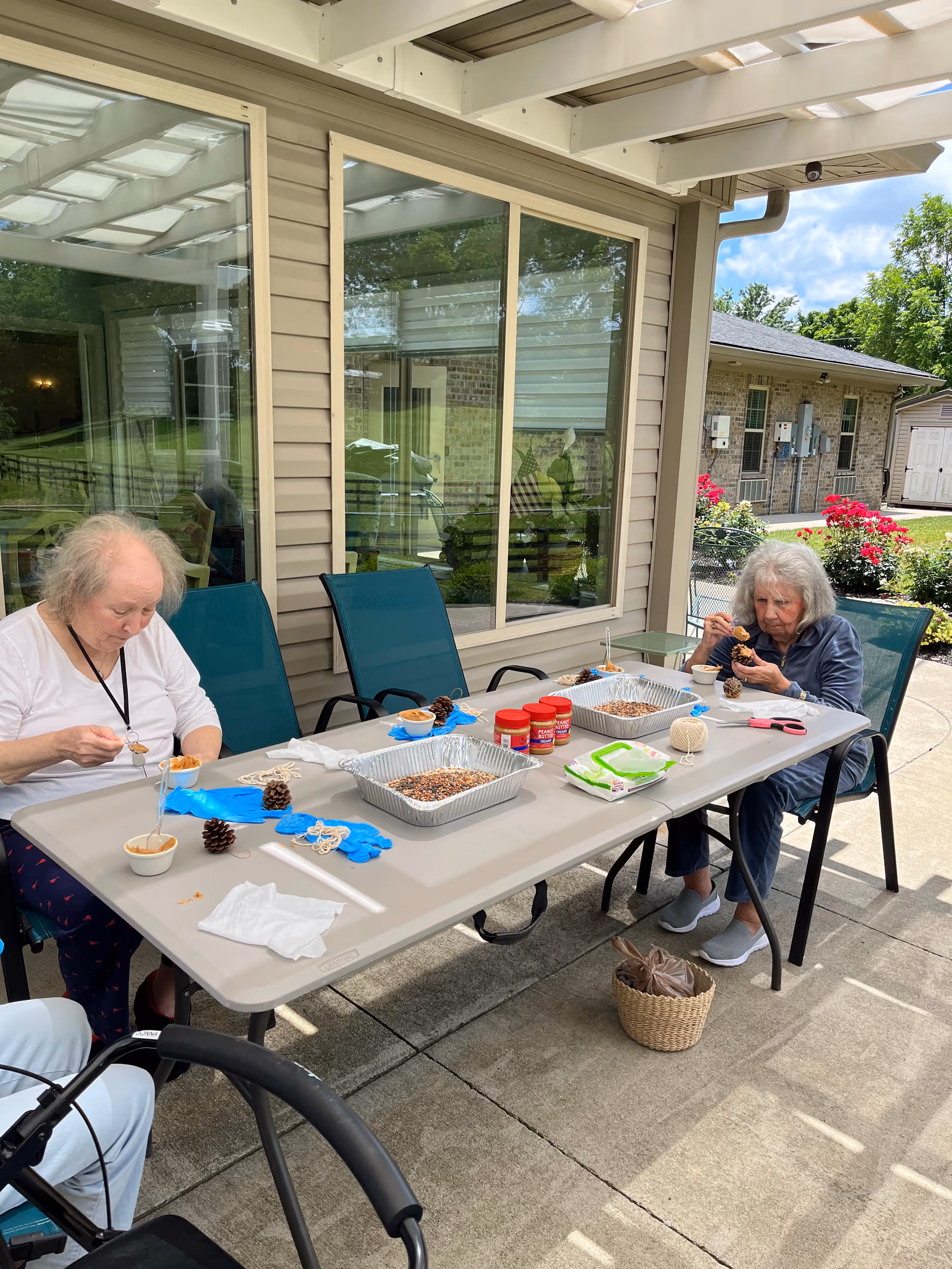 Two elderly women sitting at a table outdoors under a covered patio, engaging in a craft activity involving pine cones, peanut butter, and birdseed. The table has trays with birdseed, jars of peanut butter, string, gloves, and other craft supplies. The background shows a building with windows and a garden with flowers.