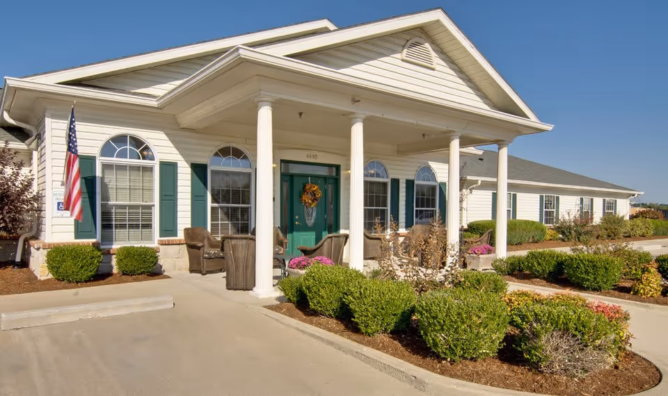 Exterior front view of a single-story senior living facility building with white siding, green shutters, and a green front door decorated with a wreath. The entrance has a covered porch supported by white columns, outdoor seating with wicker chairs, and well-maintained landscaping with bushes and flowers. An American flag is displayed near the entrance under a clear blue sky.