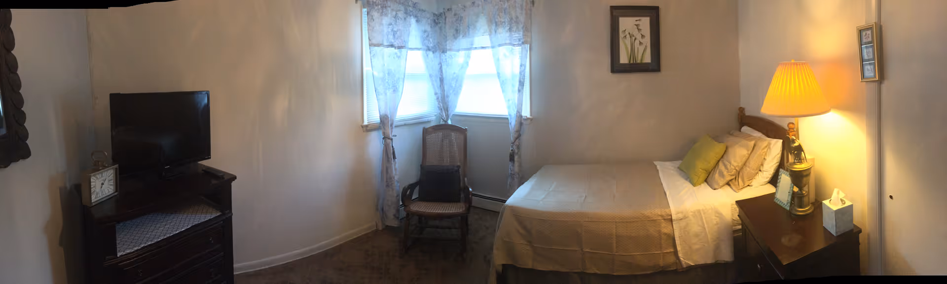 Pelican Hill facility bedroom with bed, dresser, nightstand lamp, and window with sheer curtains.