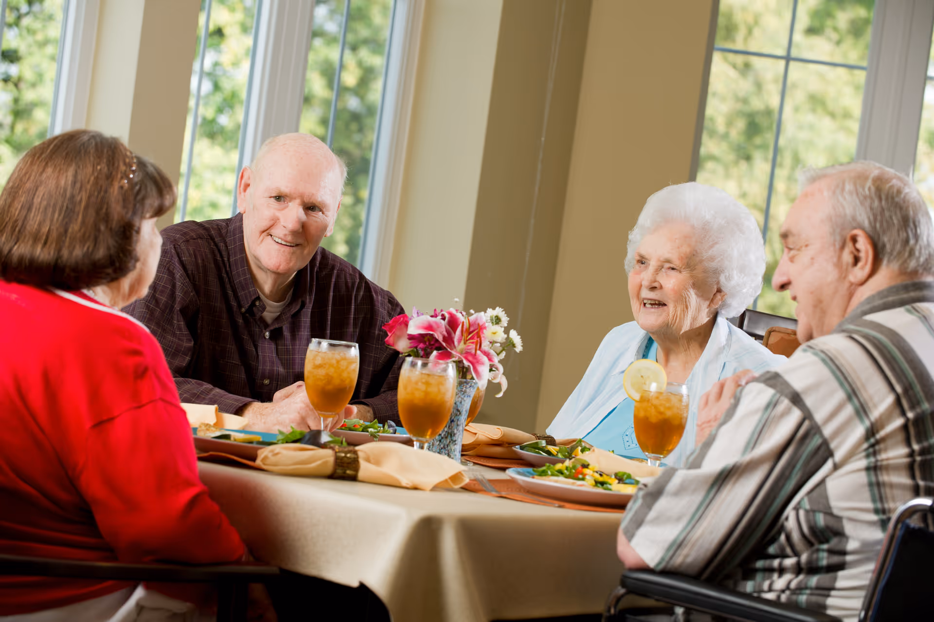 Four elderly people sitting around a dining table in a well-lit room with large windows, enjoying a meal and drinks while smiling and conversing. The table is set with plates of food, glasses of iced tea with lemon, napkins, and a vase with flowers.