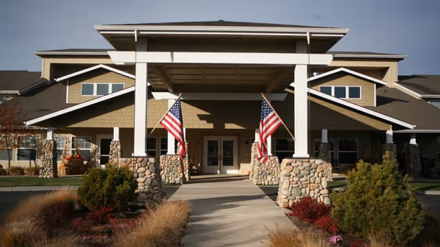 Front exterior view of Prestige Senior Living High Desert building with a covered entrance supported by stone pillars, two American flags on either side of the entrance, and landscaped bushes and grasses along the walkway.