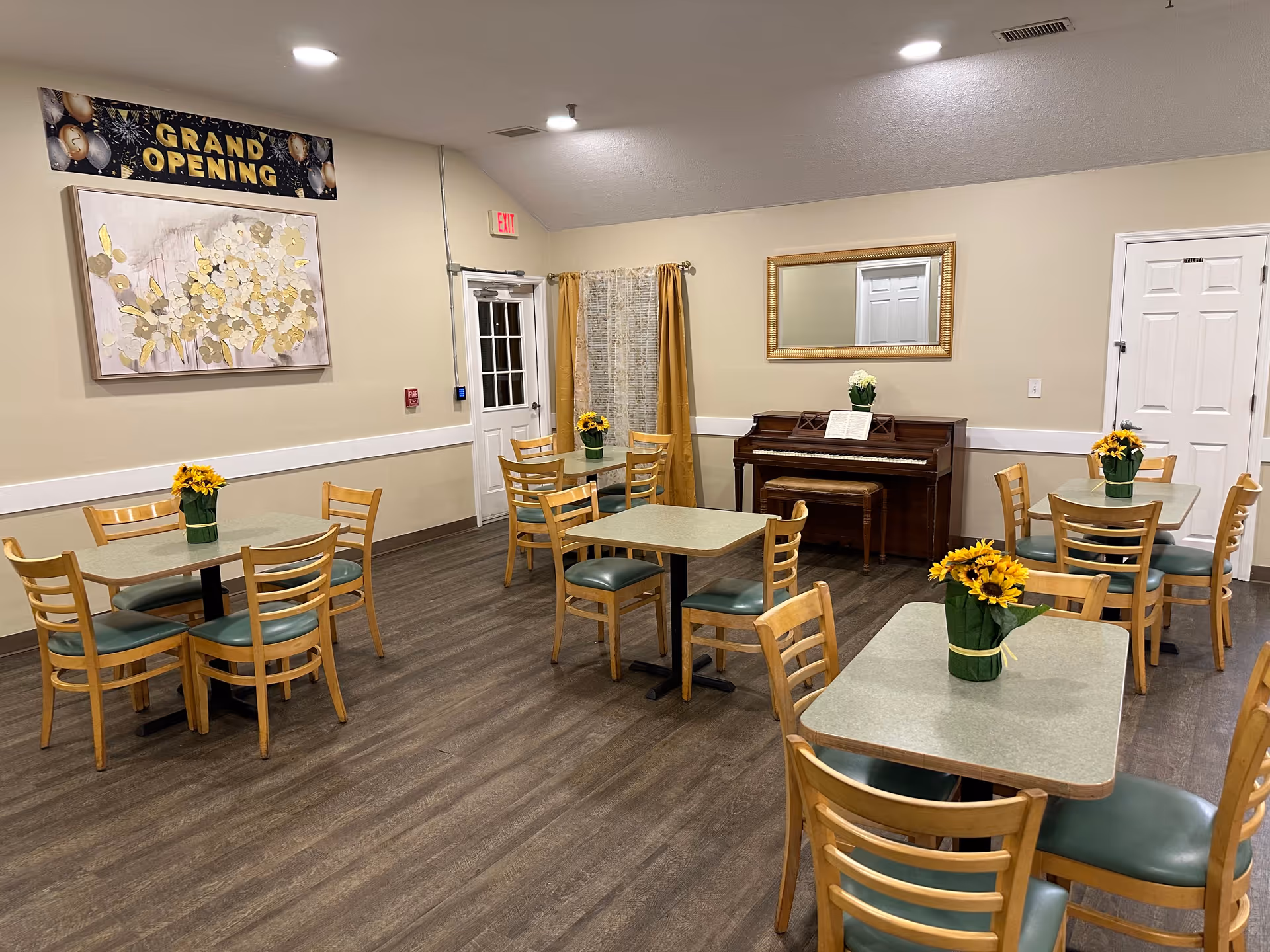 Interior of a dining room with several tables and wooden chairs with green cushions. Each table has a small vase with yellow flowers. There is a piano against the wall with a mirror above it, and a 'Grand Opening' banner is displayed on the wall. The room has wood flooring and light-colored walls.