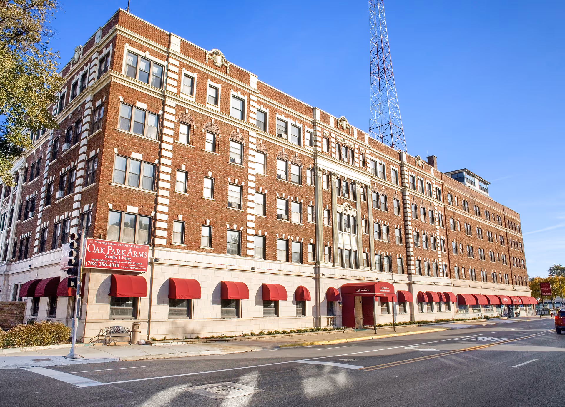 Large brick multi-story senior living building labeled Oak Park Arms with red awnings along the sidewalk under a clear blue sky.