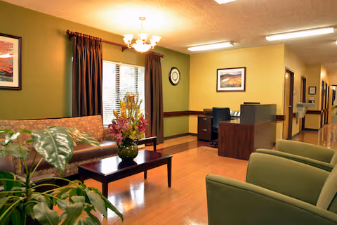 Cozy senior living lounge with green chairs, a sofa, plants, and a wooden floor.