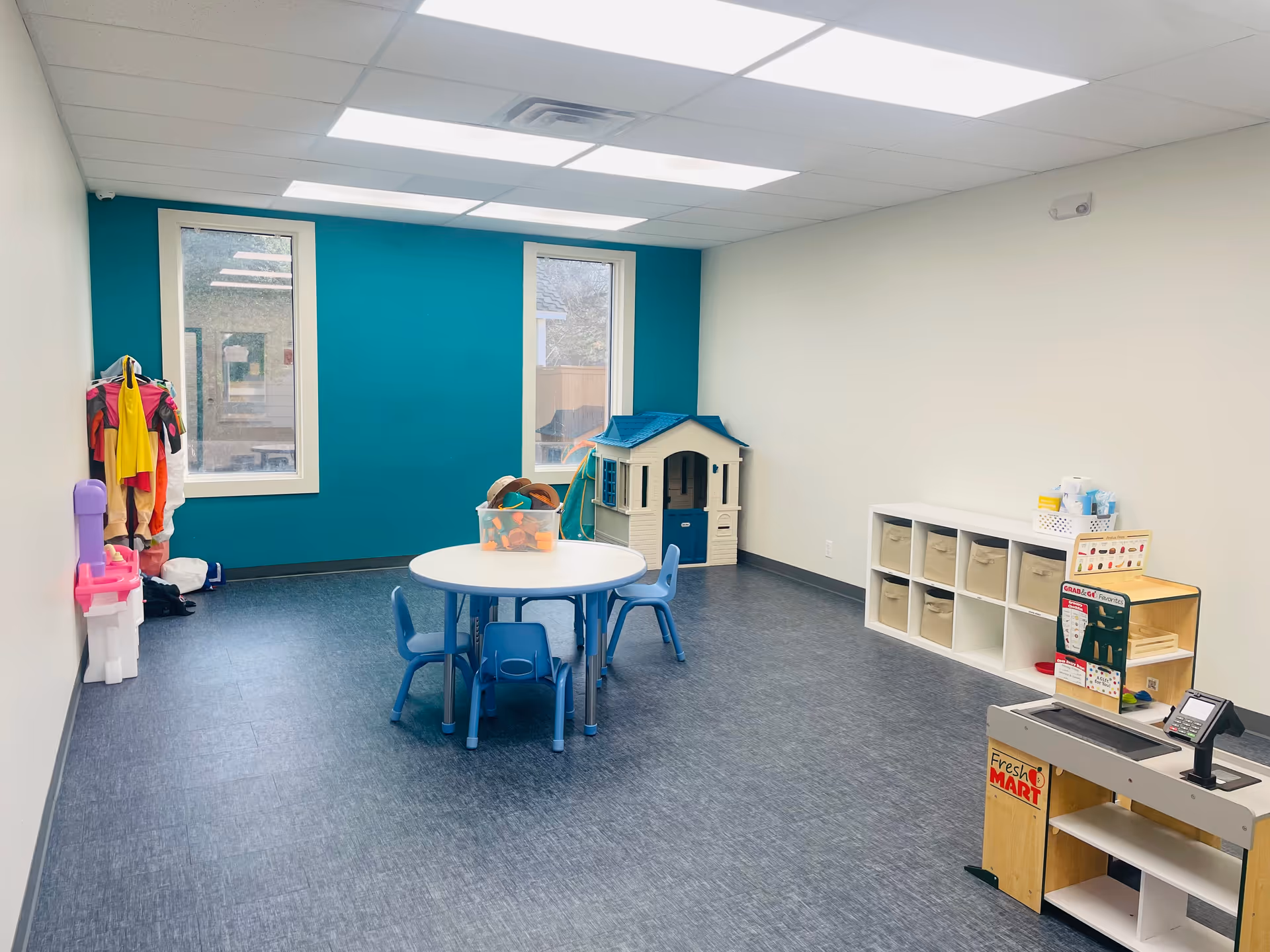 Bright interior playroom with a round table, blue chairs, cubbies, and a toy house.