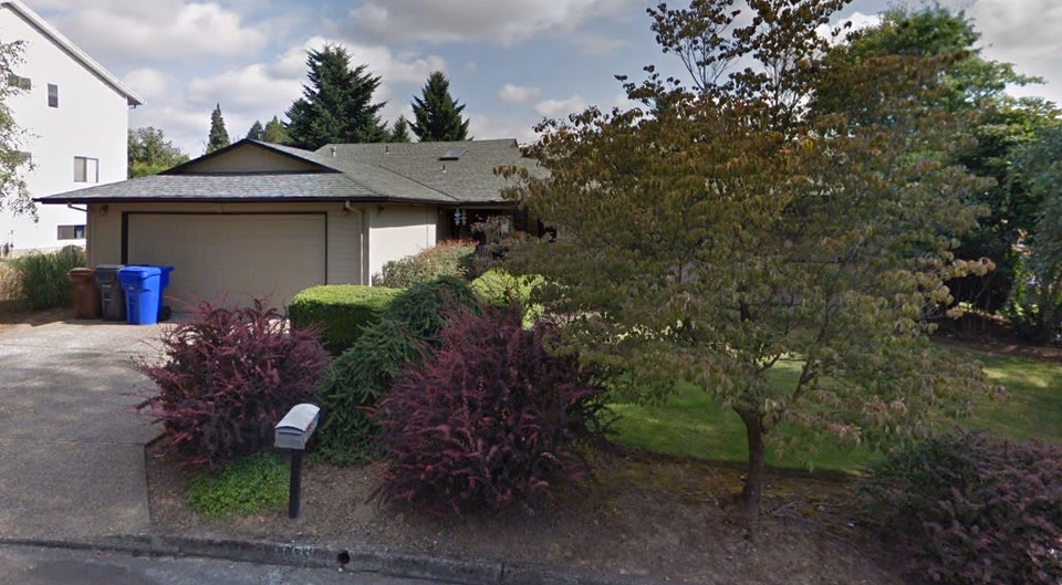 Front exterior of a single-story house with a two-car garage, driveway, mailbox, and landscaping with shrubs and trees.